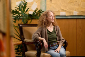 A person with curly hair sits in an ornate chair, gazing thoughtfully to the side in a warmly lit room with a large plant and a bulletin board in the background.